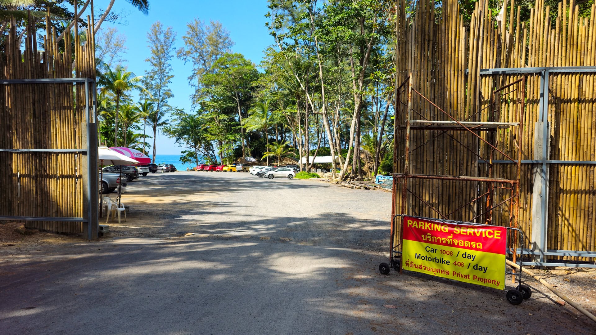 Dirt parking area near entrance with bamboo fencing and view of sea beyond trees.