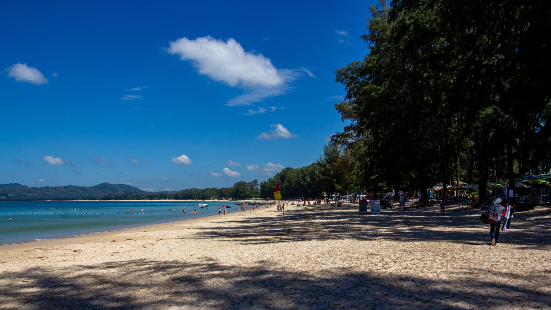 View north along Bang Tao Beach with shaded casuarina trees and swimmers in calm water.