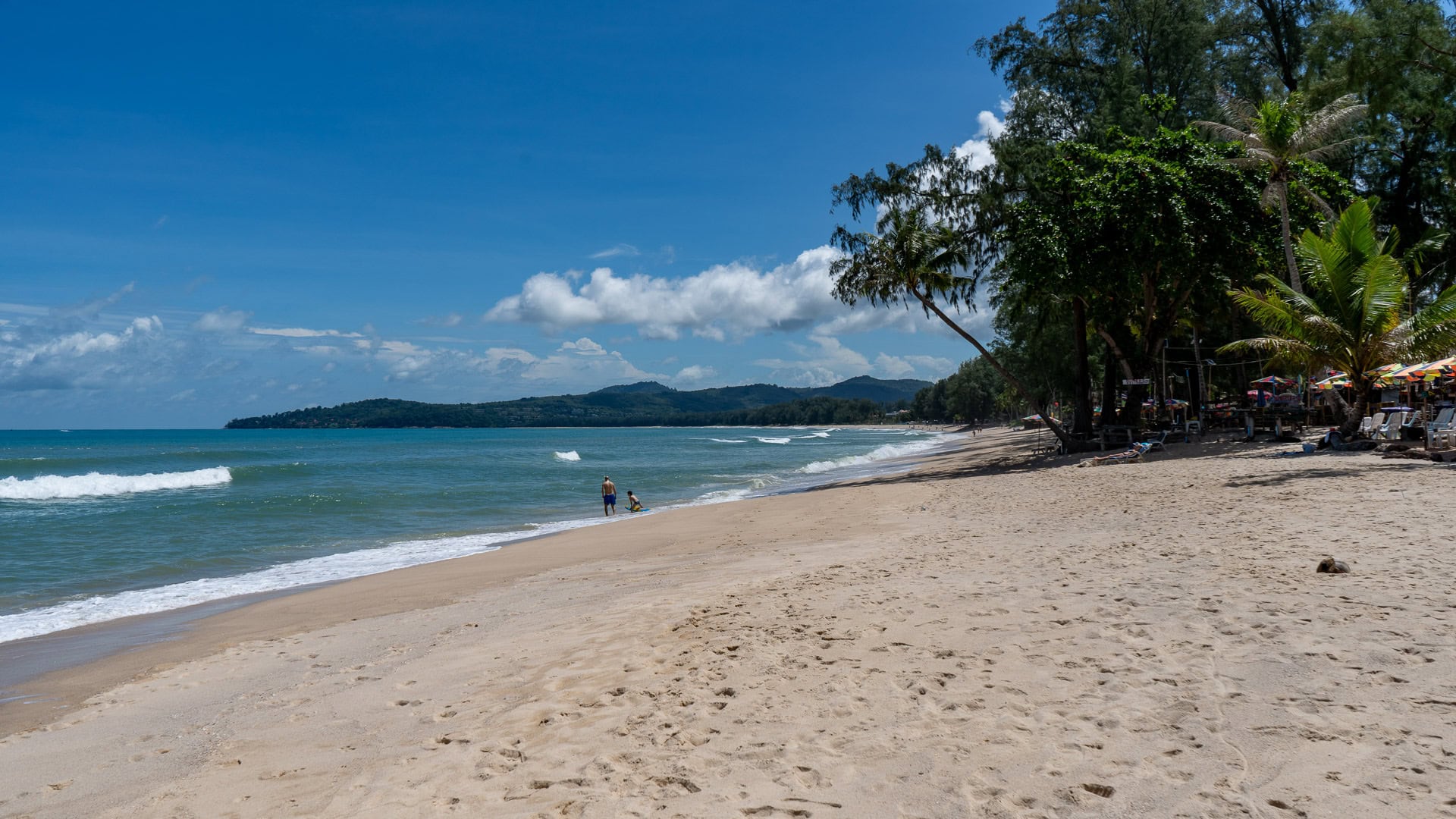 Moderate waves reaching the shoreline with beachside trees and umbrellas.