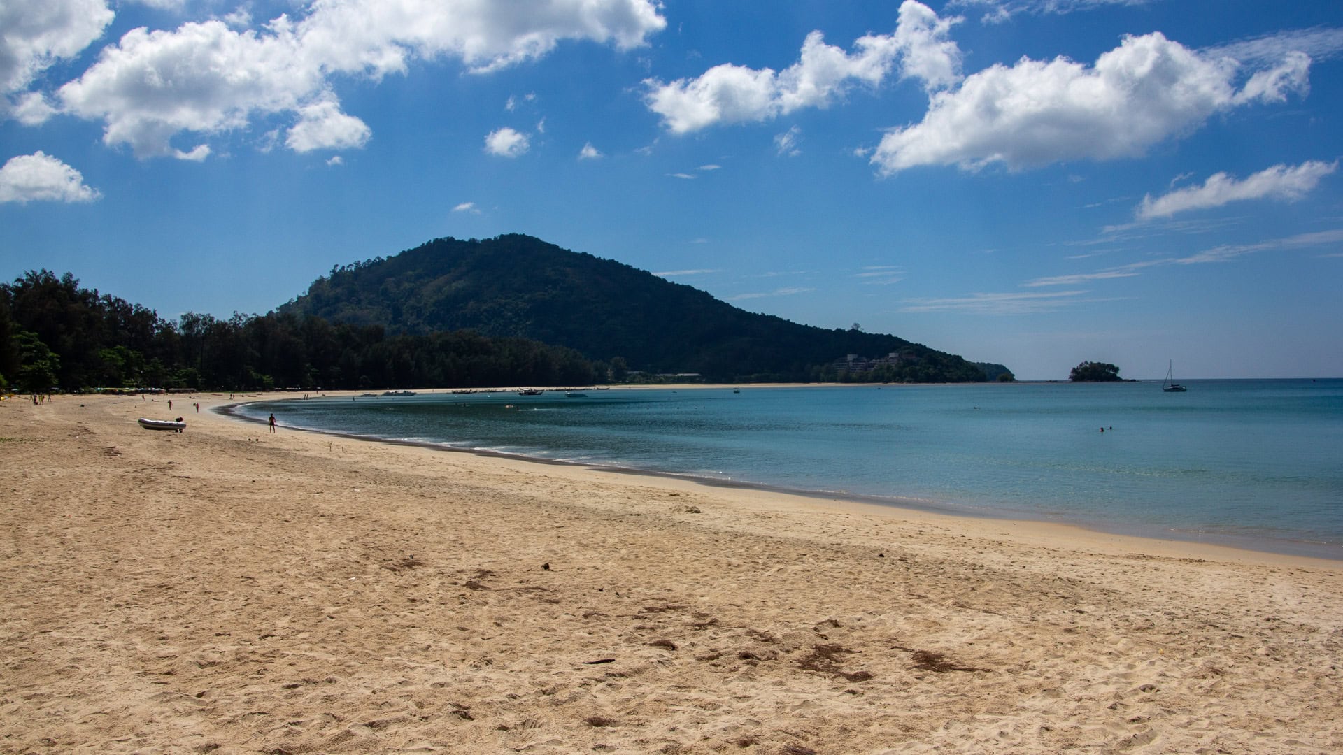 Calm blue water and wide sandy shoreline at Nai Yang Beach Phuket, with tree shade along the coast.