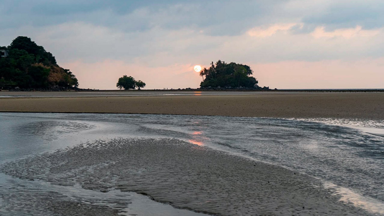 Man standing in shallow water at Nai Yang Beach Phuket with distant coastline and calm sea.