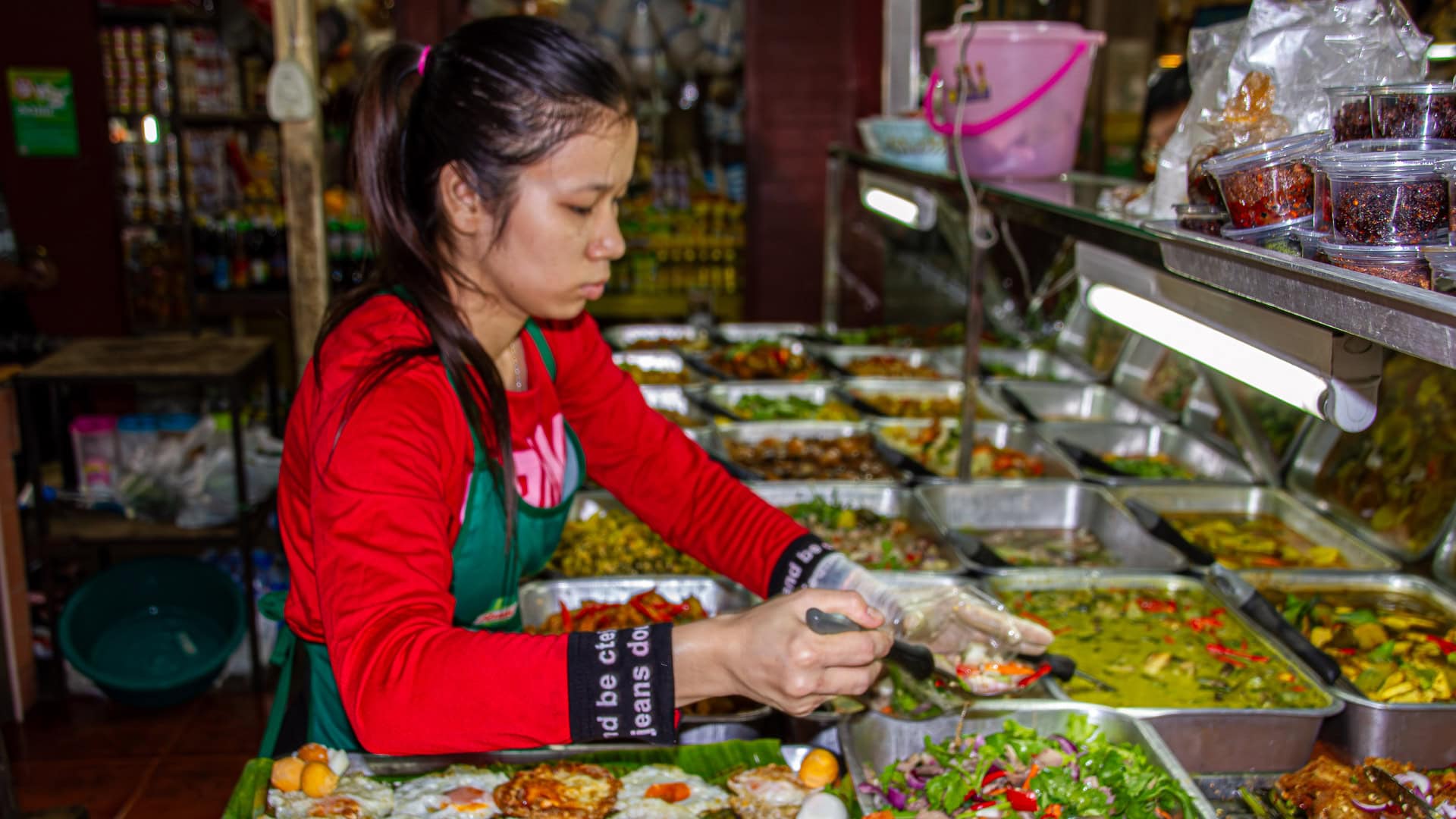 Banzaan Fresh Market Phuket prepared food stall with a vendor serving cooked dishes from metal trays.