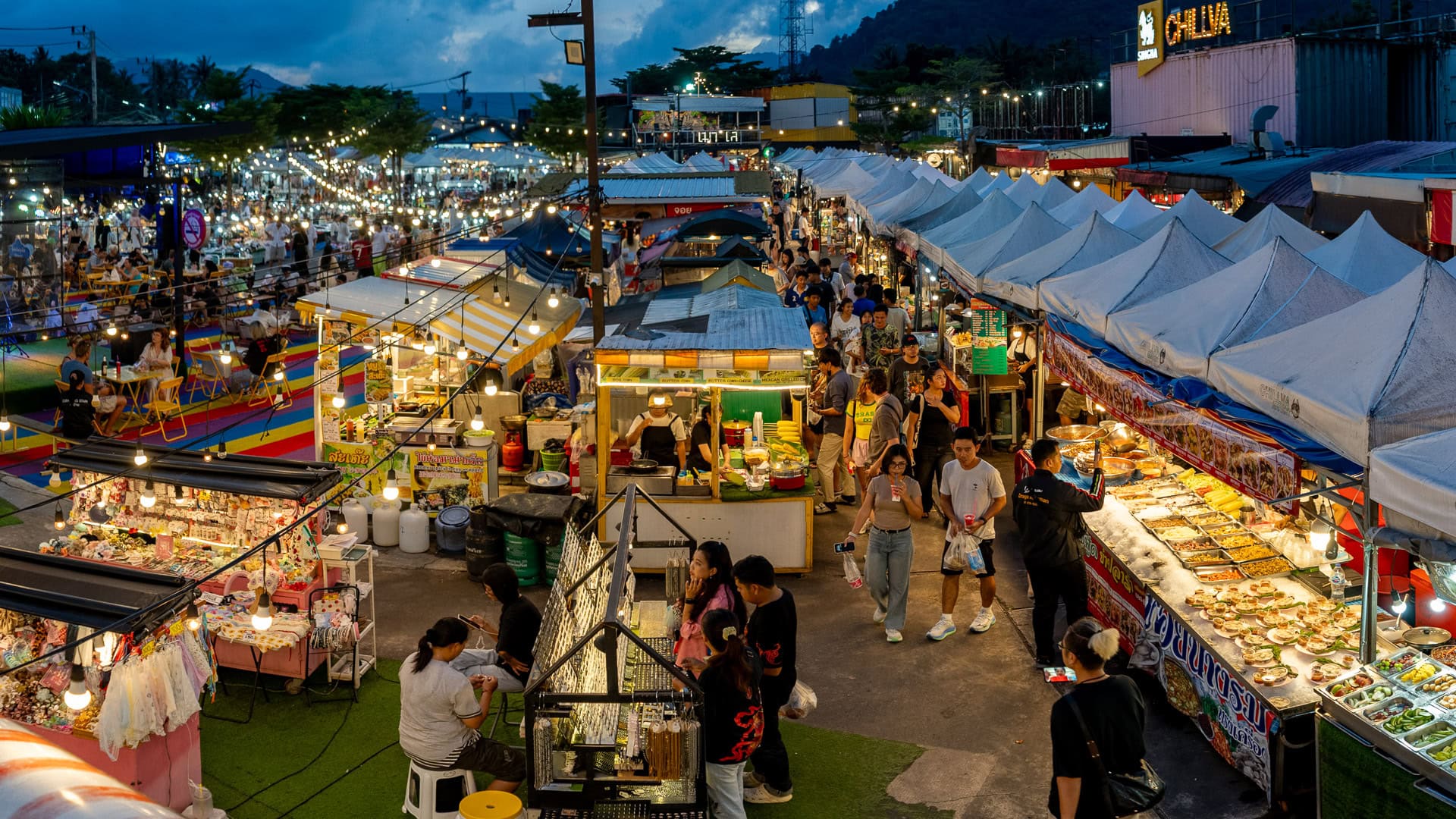 Chillva Market Phuket with multiple food stalls, prepared dishes on display, vendors serving customers, and people walking and queuing between aisles.