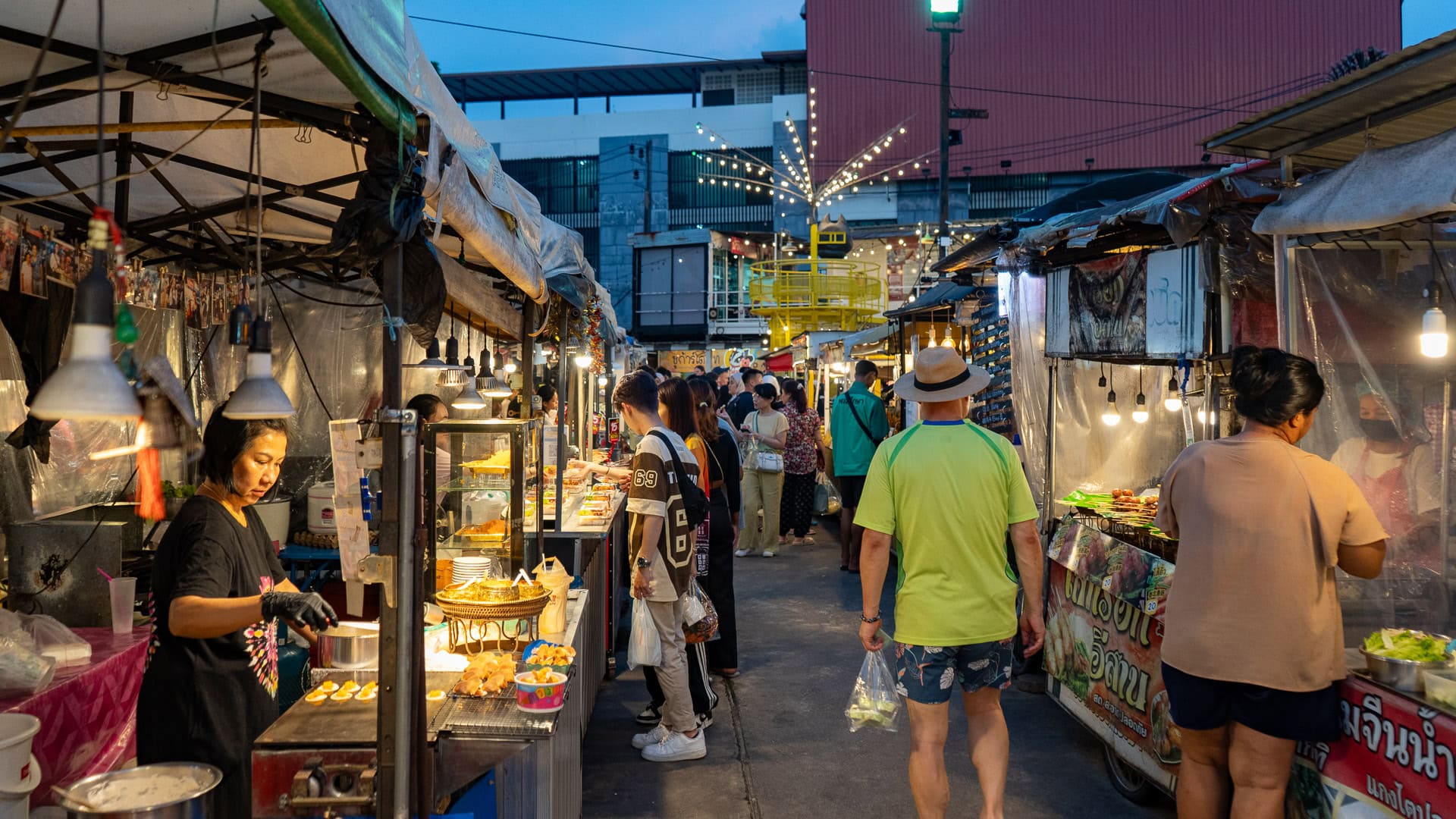 Chillva Market Phuket with food stalls along a narrow aisle, vendors cooking and serving dishes, and customers walking and stopping to choose food.