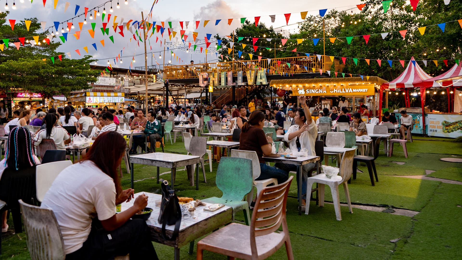 Chillva Market Phuket seating area with tables and chairs, people eating and drinking, food stalls in the background, and overhead lights and flags above the space.