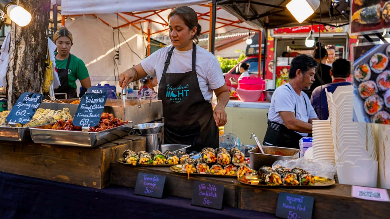 Fun Friday Boat Avenue Market Phuket food stall with a vendor preparing skewered food at a counter, cooked items arranged on trays with price signs visible
