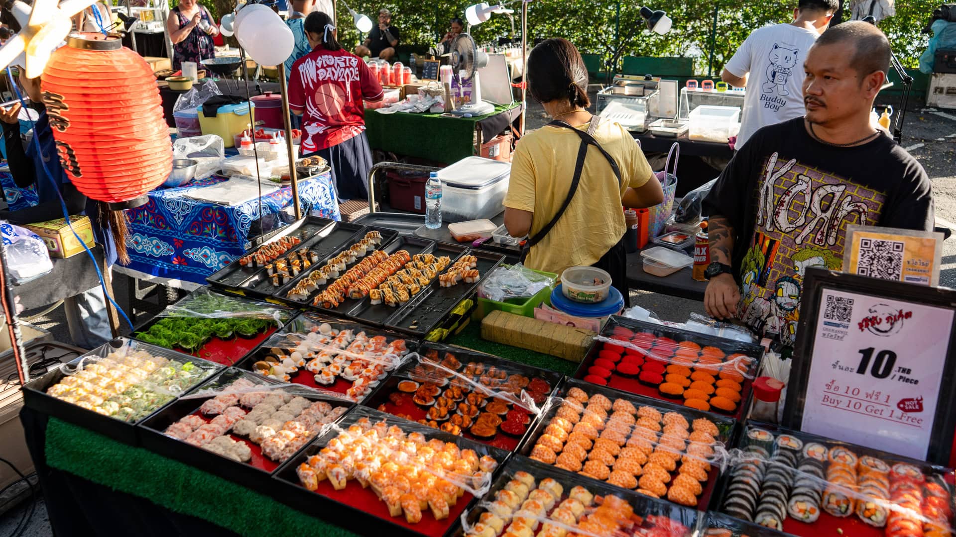 Fun Friday Boat Avenue Market Phuket food stall with trays of sushi rolls, skewered snacks, and prepared bites arranged on tables, with a vendor standing beside the display and customers nearby