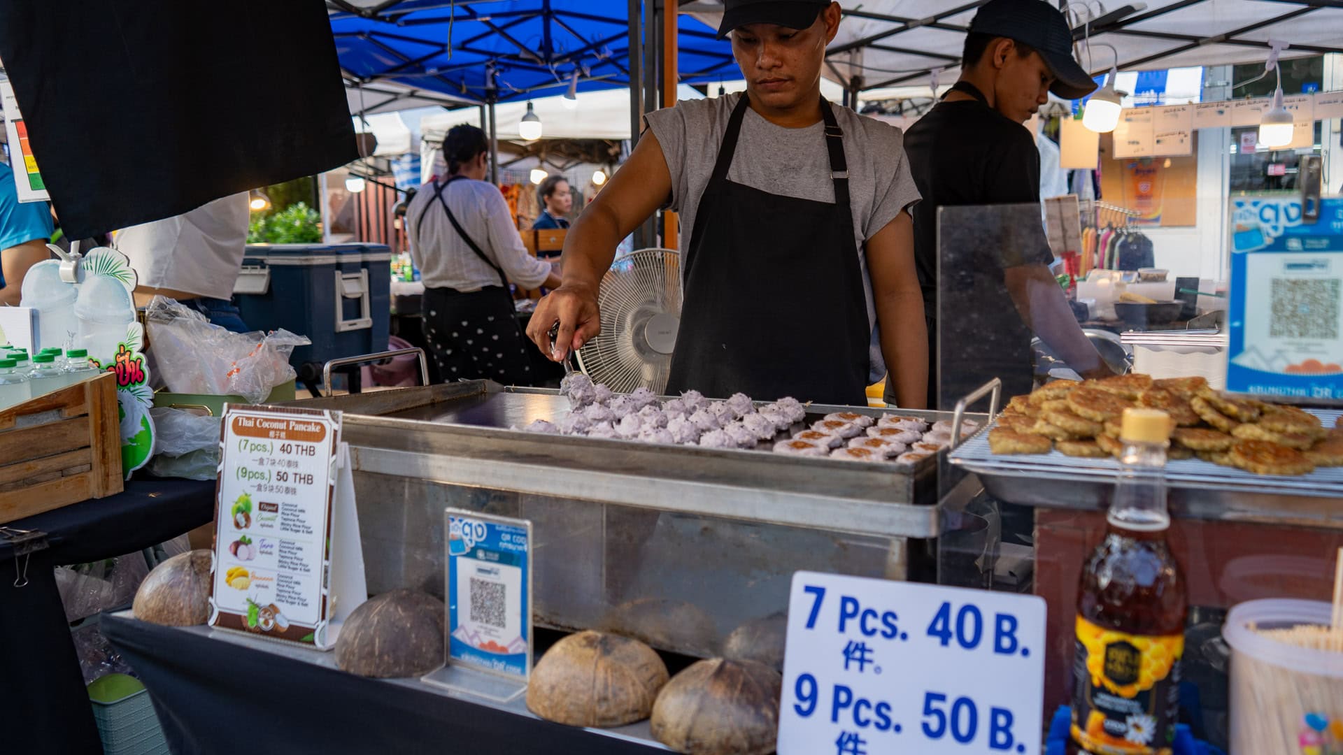 Fun Friday Boat Avenue Market Phuket coconut pancake stall with a vendor cooking batter on a flat metal grill, finished pancakes stacked beside the cooking surface and price signs visible
