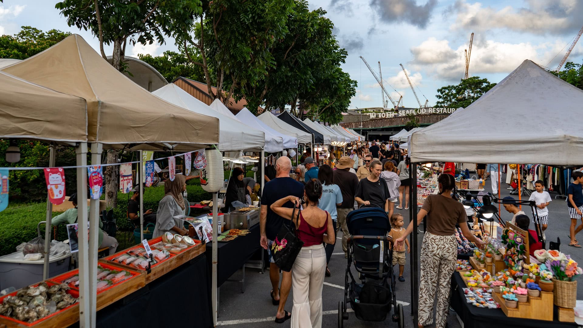 Fun Friday Boat Avenue Market Phuket main aisle with rows of white tents, food and craft stalls on both sides, and people walking, browsing, and pushing a stroller through the market walkway