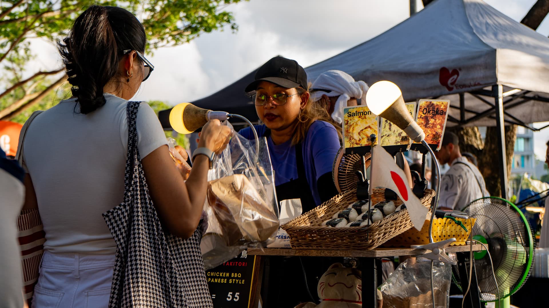 Fun Friday Boat Avenue Market Phuket food stall with a vendor handing a takeaway bag to a customer across the counter, prepared food displayed on the table and market tents visible behind the stall