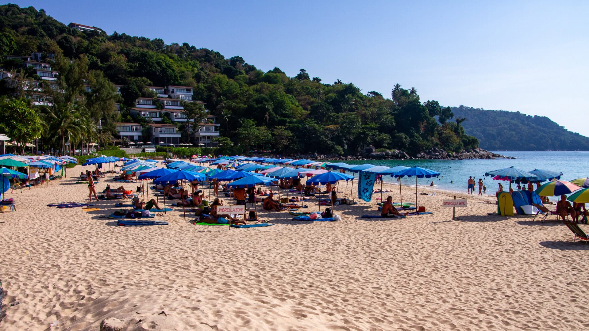 Kata Noi Beach Phuket crowded sunbed area with blue umbrellas and hillside buildings