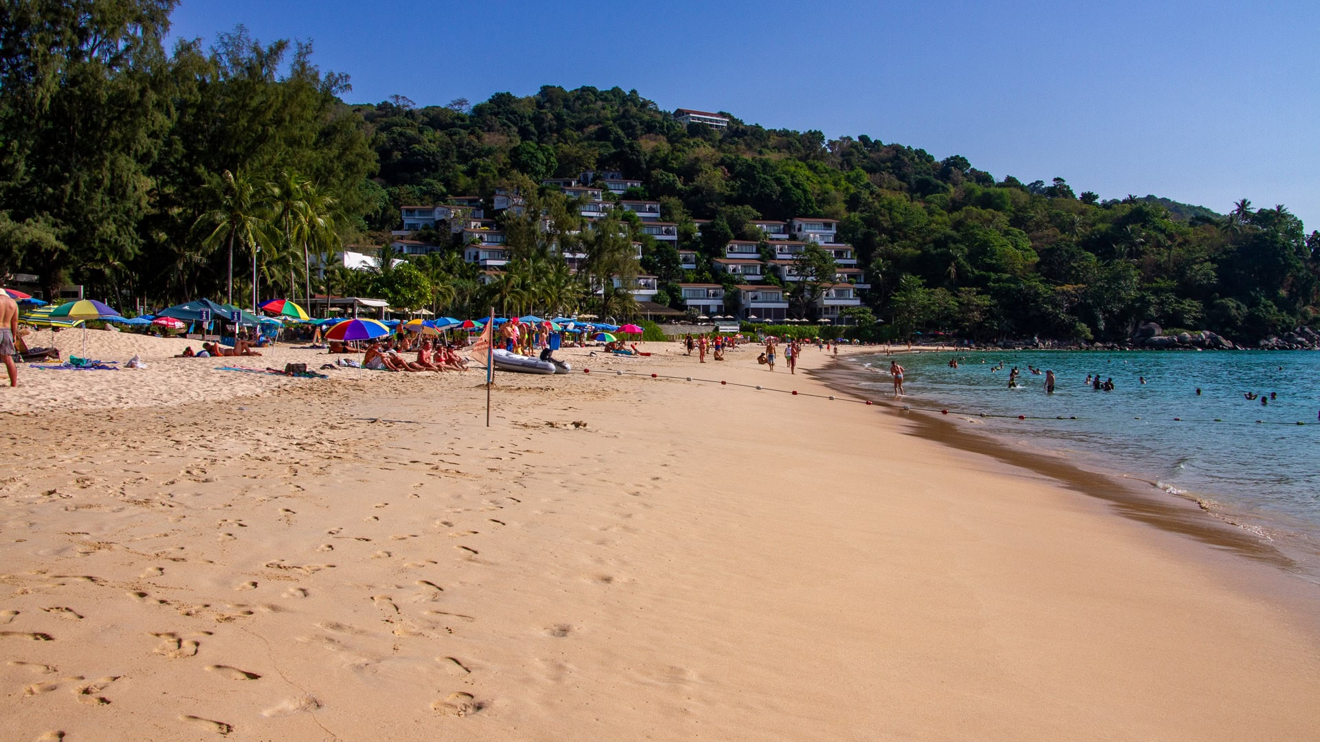 Kata Noi Beach Phuket shoreline with swimmers, umbrellas, and hillside resort buildings