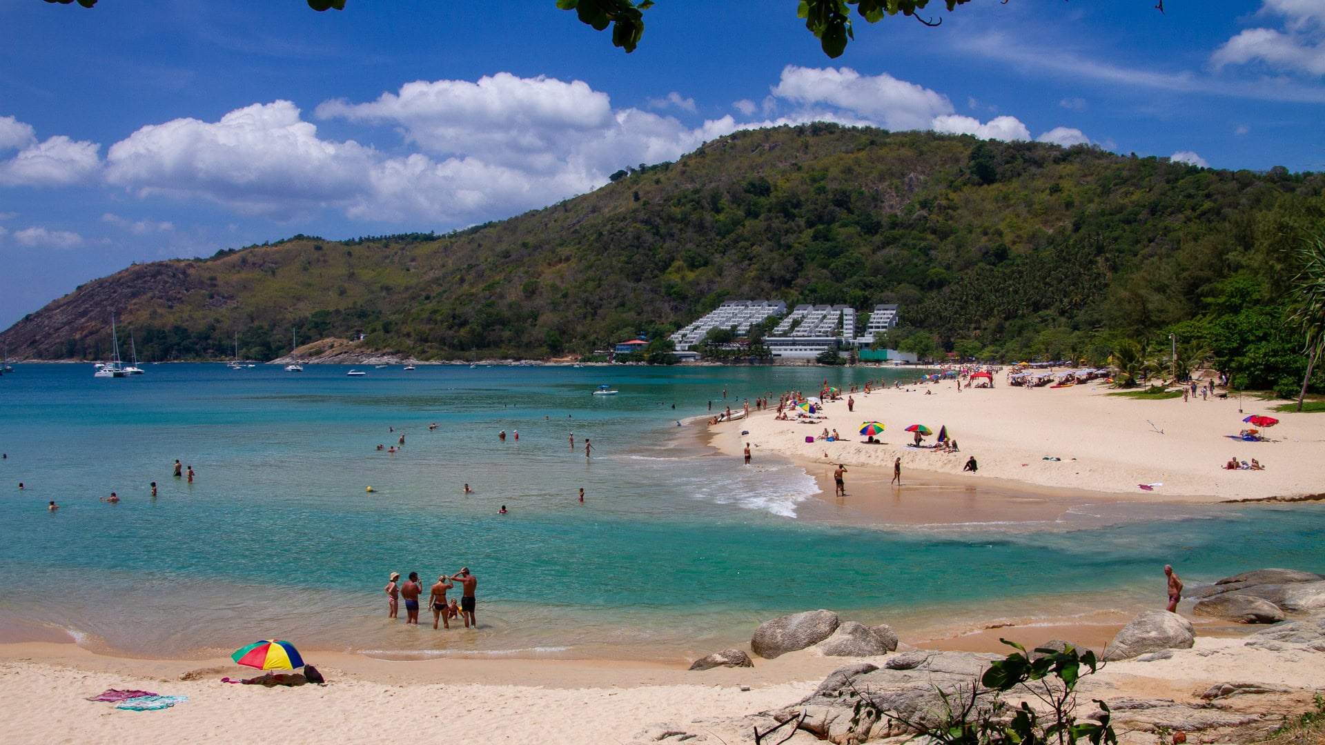 Nai Harn Beach Phuket central bay with umbrellas, swimmers, and anchored boats offshore