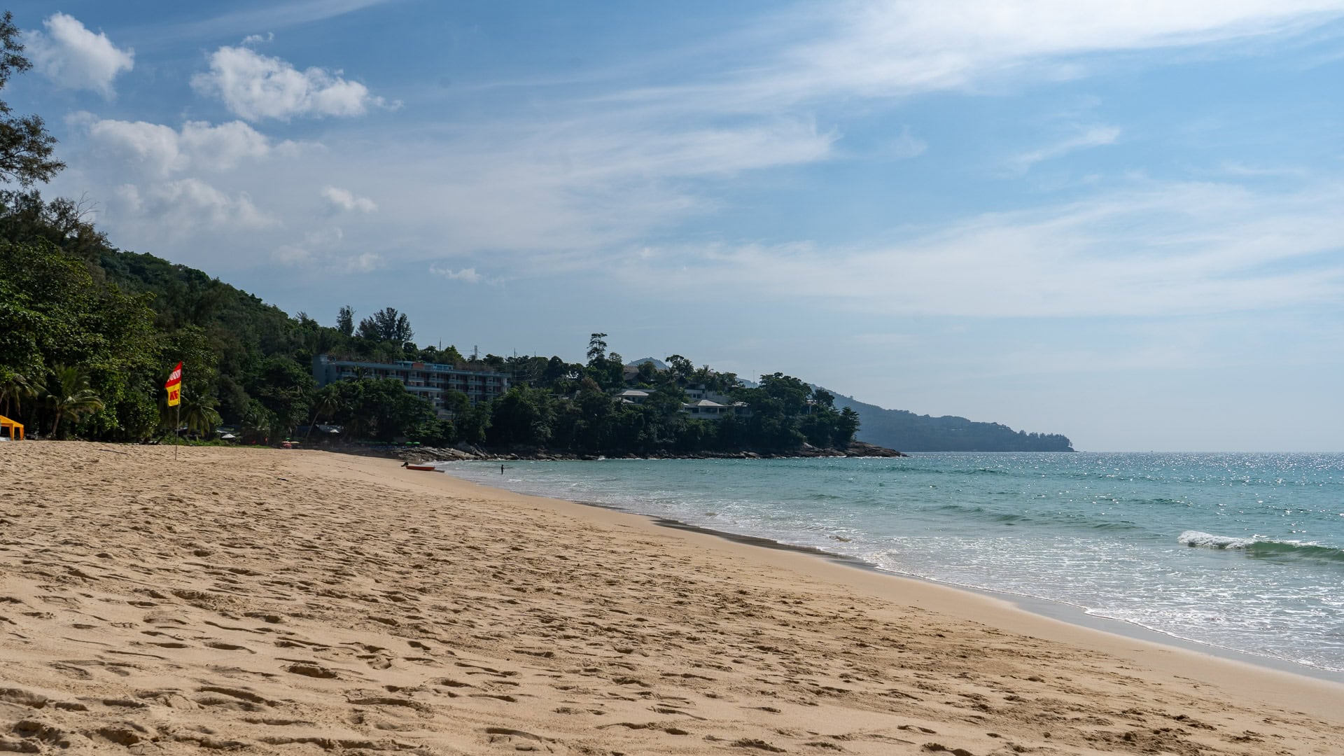 Wide sandy shoreline with calm turquoise water seen from shaded trees.