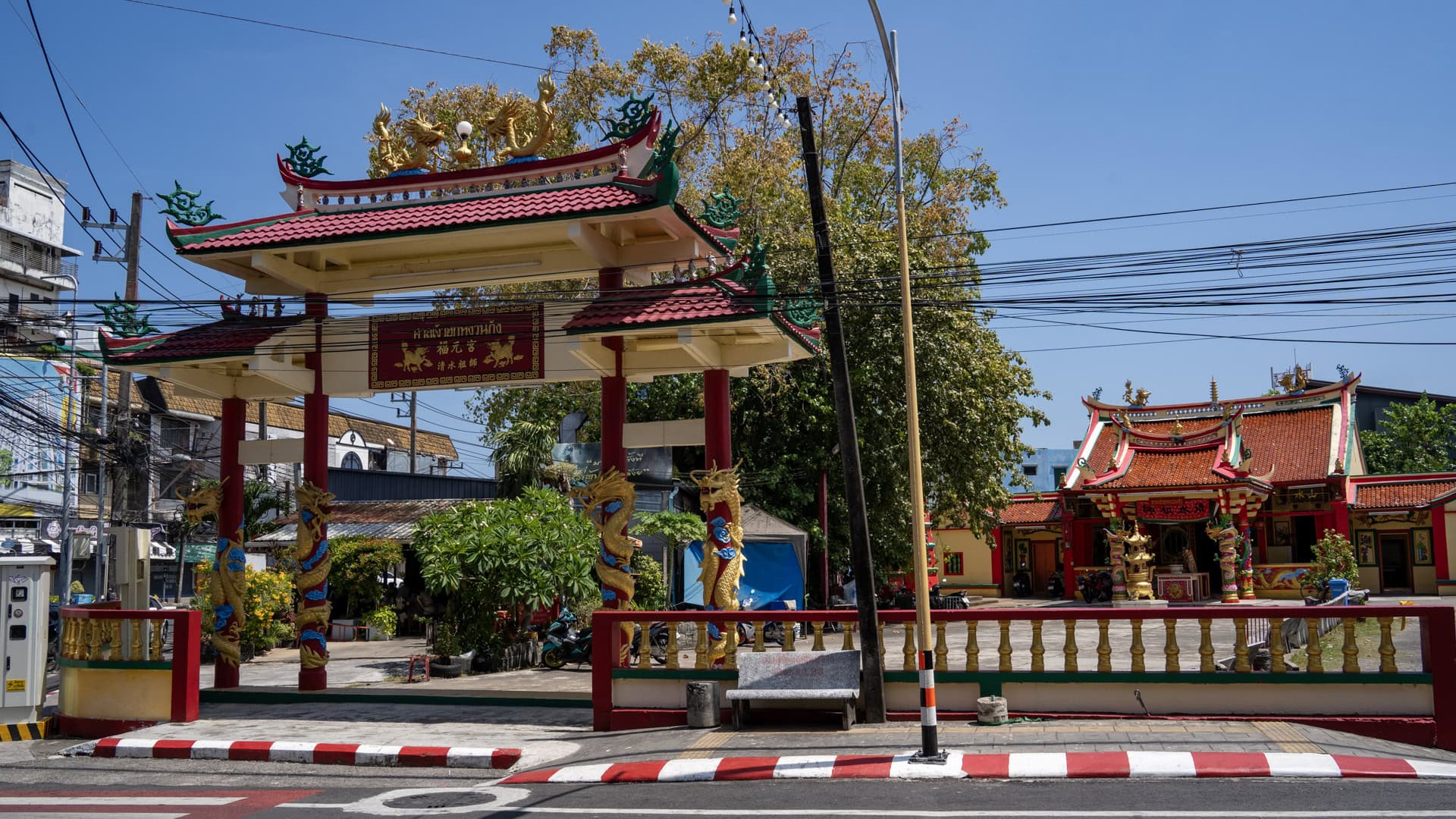 A Chinese shrine gate with red pillars, green roof ornaments, and dragon-wrapped columns stands beside a busy road, with the main shrine hall visible behind it.