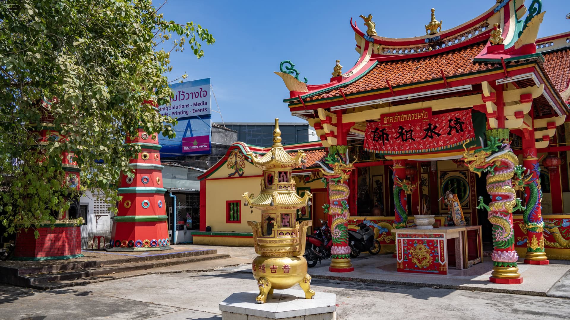A gold incense burner stands in the forecourt before a red-and-gold Chinese shrine, with dragon-wrapped entrance columns and a tall red pagoda-like tower at the left.