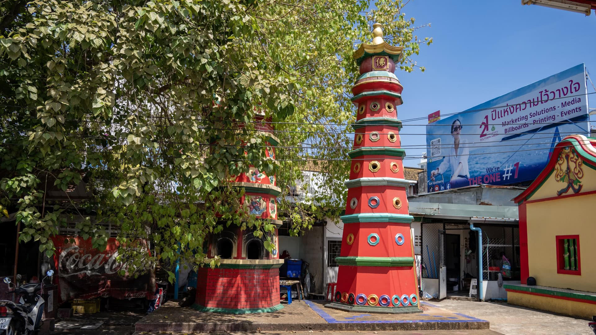 Two red pagoda-like towers stand under a leafy tree beside a Chinese shrine building, with overhead wires and a roadside billboard in the background.