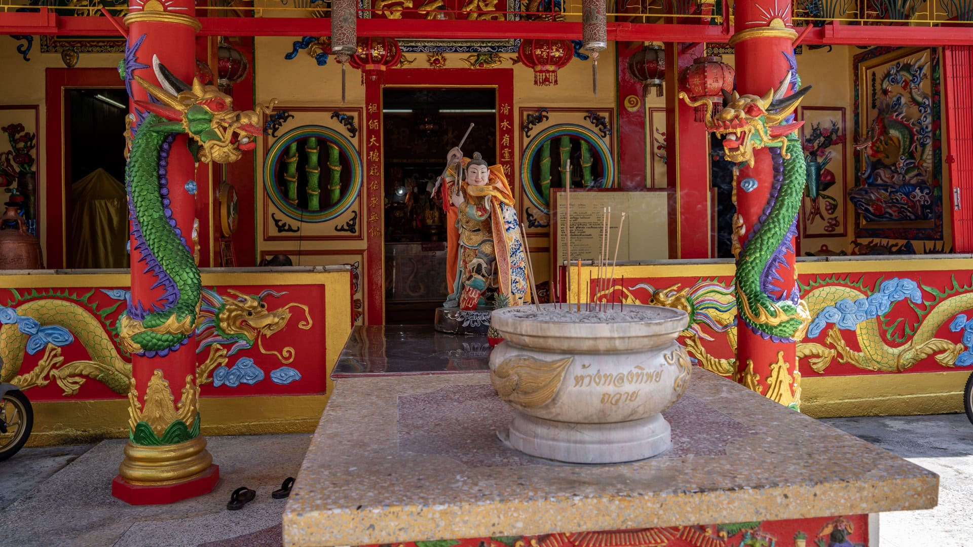 A stone incense urn sits on a terrazzo table before a Chinese shrine entrance, flanked by red columns wrapped with green dragons and a standing deity figure inside.