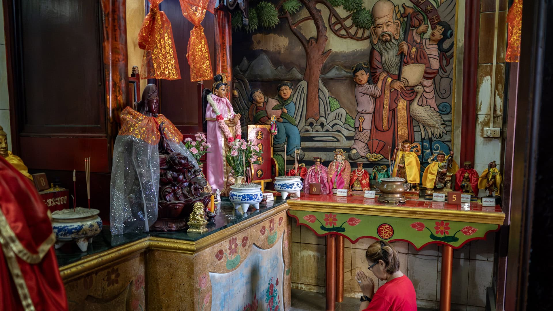 A side altar inside the shrine holds small robed figures, incense pots, and flower offerings, with a painted wall panel of an elderly figure and children behind.