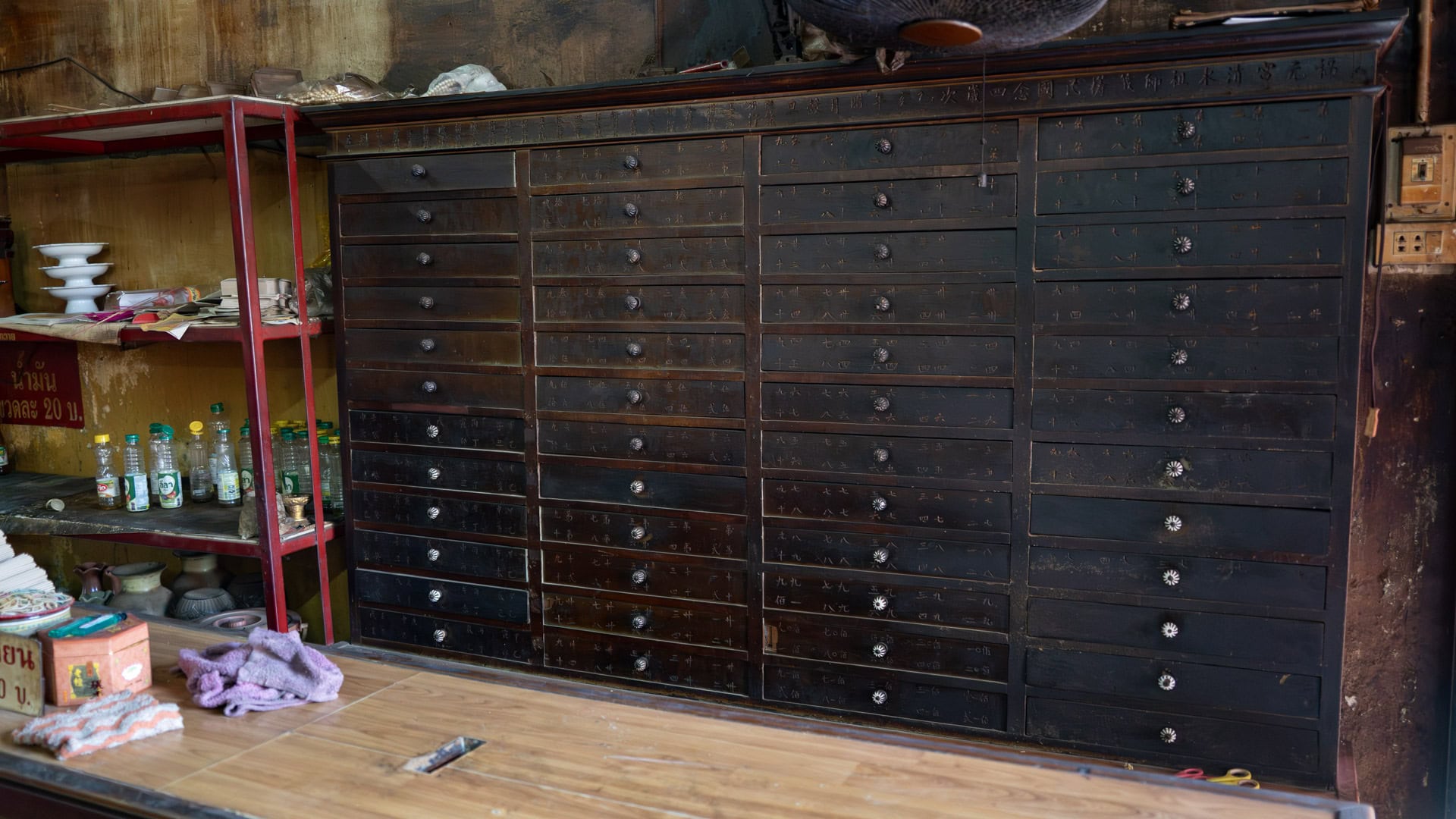 A large dark wooden cabinet with many narrow labeled drawers stands against an interior wall, beside a red metal shelf holding bottles and dishes.