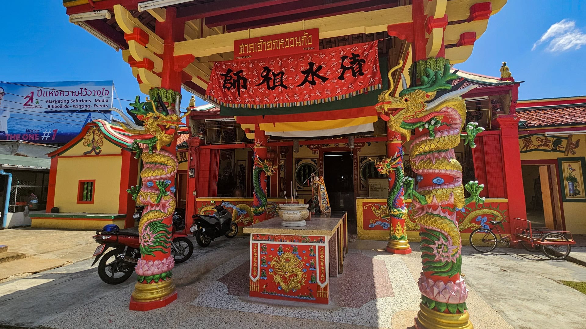 The front porch of a Chinese shrine has two brightly painted dragon-wrapped columns, a red hanging banner, and a central incense table, with motorcycles parked at the side.