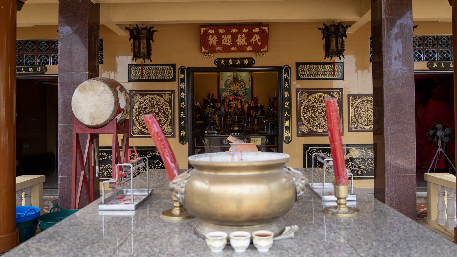 A brass incense burner sits on a stone table in front of the shrine hall, with red candles, a drum, and altar figures visible inside.