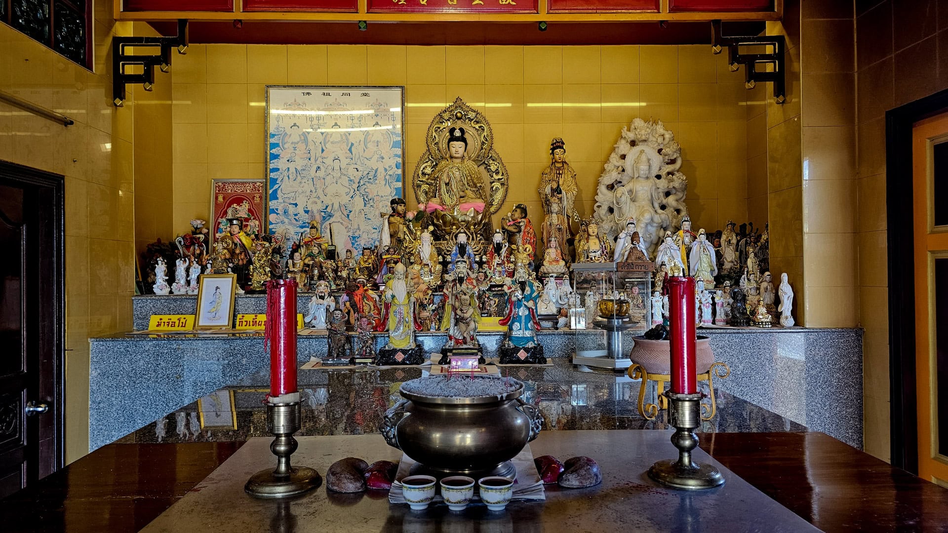 A side altar holds many small deity figures, with red candles and a black incense burner in front of a gold-seated figure and white statues.